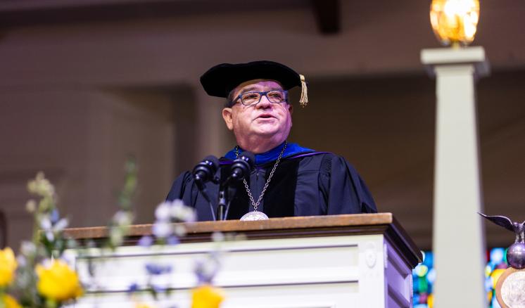 Person in academic regalia stands at a podium with floral arrangements.
