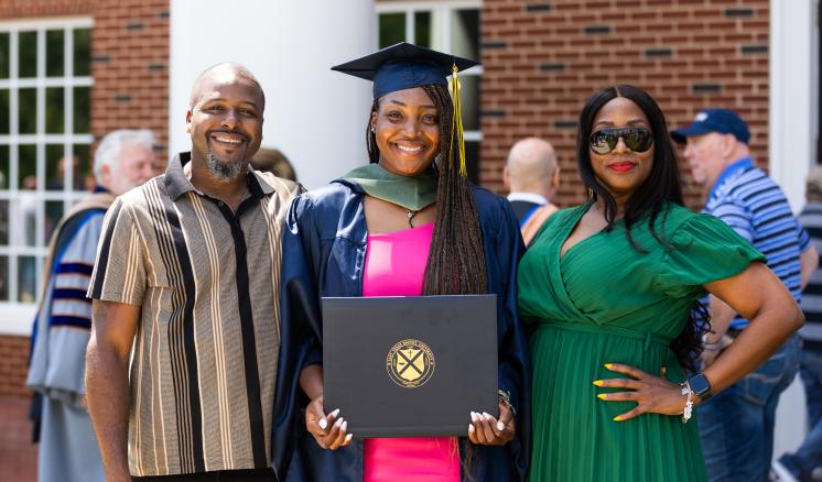 Three people, one in graduation attire, standing and smiling outside a brick building.
