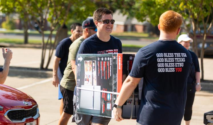 2 men carrying a large box outside in casual clothing with sunglasses