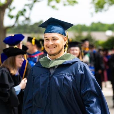Smiling graduate in a navy blue gown and cap with a green hood during a graduation ceremony.