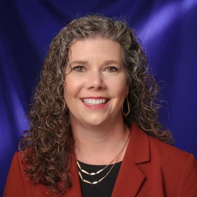 A woman wearing a red blazer with gold necklaces, and long curly hair, standing infront of a blue backdrop
