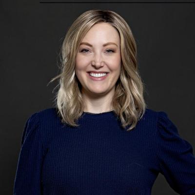 A smiling woman with shoulder-length blonde hair is wearing a dark blue long-sleeved shirt, posed against a plain dark background.