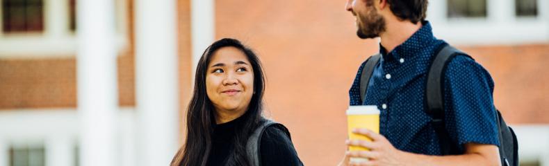 Two students walking and talking in front of a brick building on a college campus.