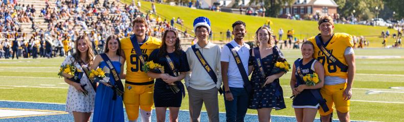 A group of students, including football players and homecoming royalty, poses together on a football field during a sunny homecoming event.