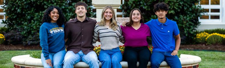 Five college students sit on a circular raised flower bed surrounding ETBU's Light on the Hill