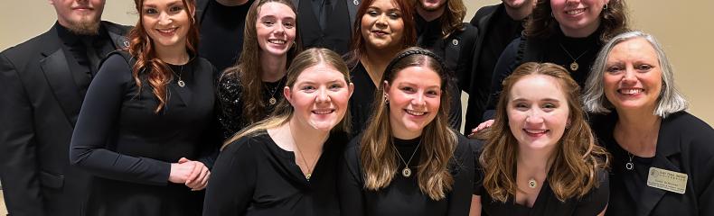 A group of thirteen people in formal black attire, smiling indoors against a beige wall.